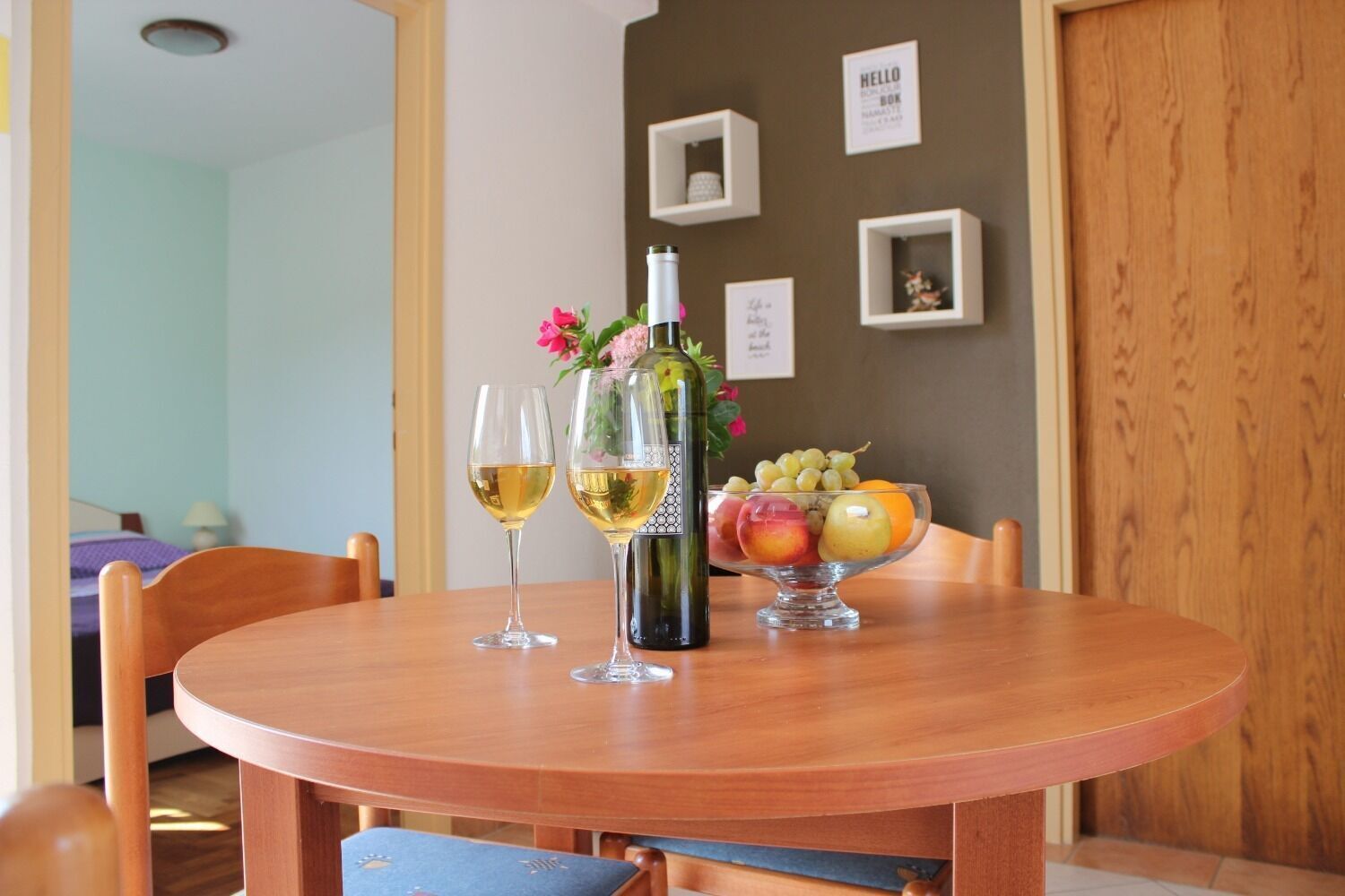 Une table de salle à manger ronde en bois, avec deux verres à vin, une bouteille et un panier de fruits.