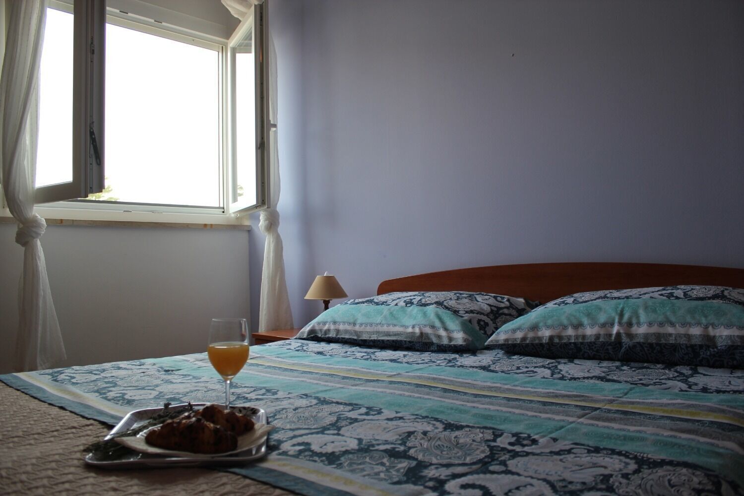 A bedroom with a bed, a bedside table, a lamp, a window with curtains, and a tray with food and a drink.