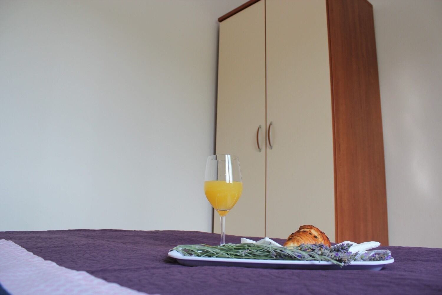A bed with a purple blanket, a glass of orange juice, a plate with food, and a croissant.