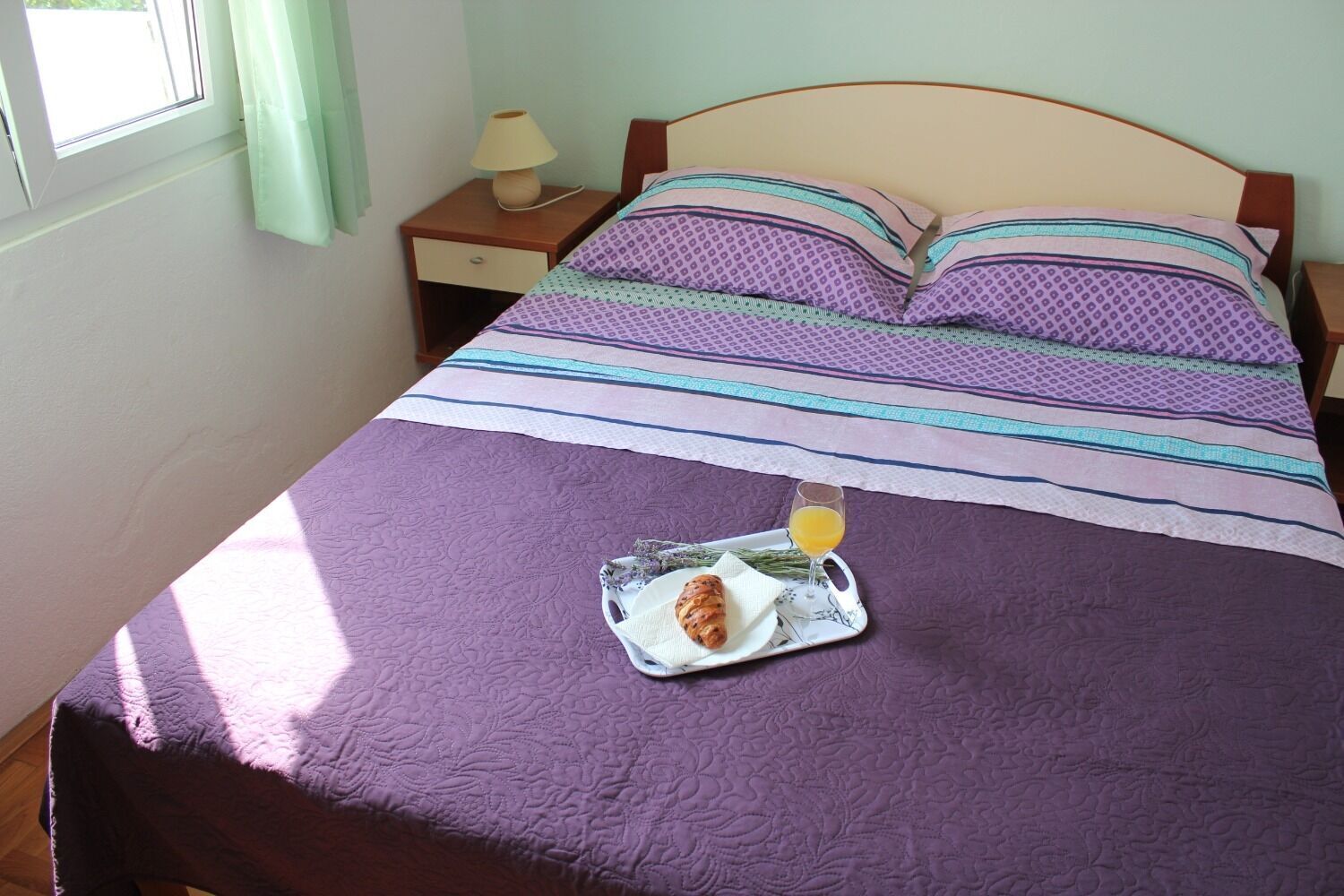 A neatly made bed with a purple quilt, a tray with croissant and orange juice, and a nightstand with a lamp.