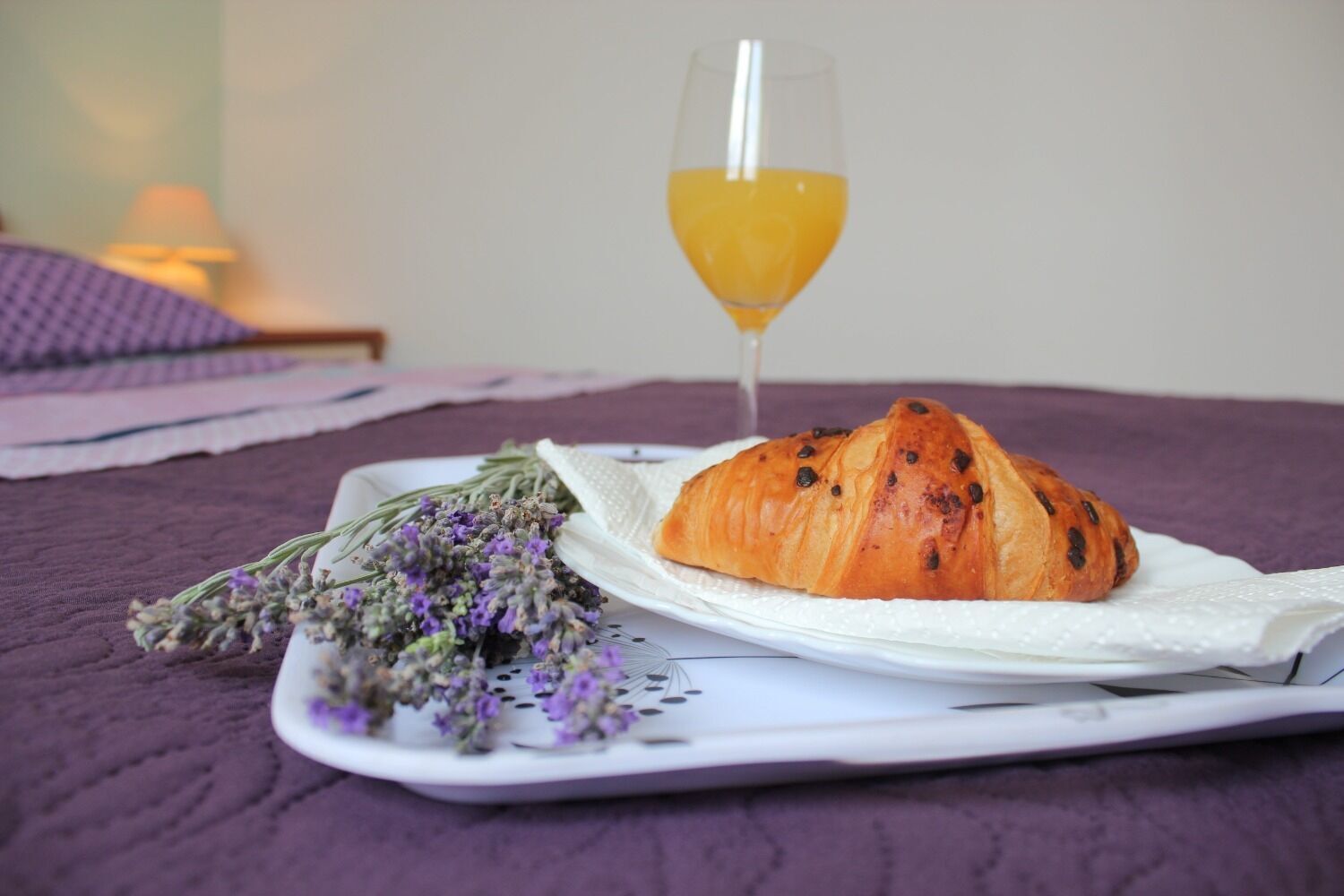 Una bandeja con un croissant, un vaso de zumo de naranja y un ramo de flores de lavanda sobre una cama.