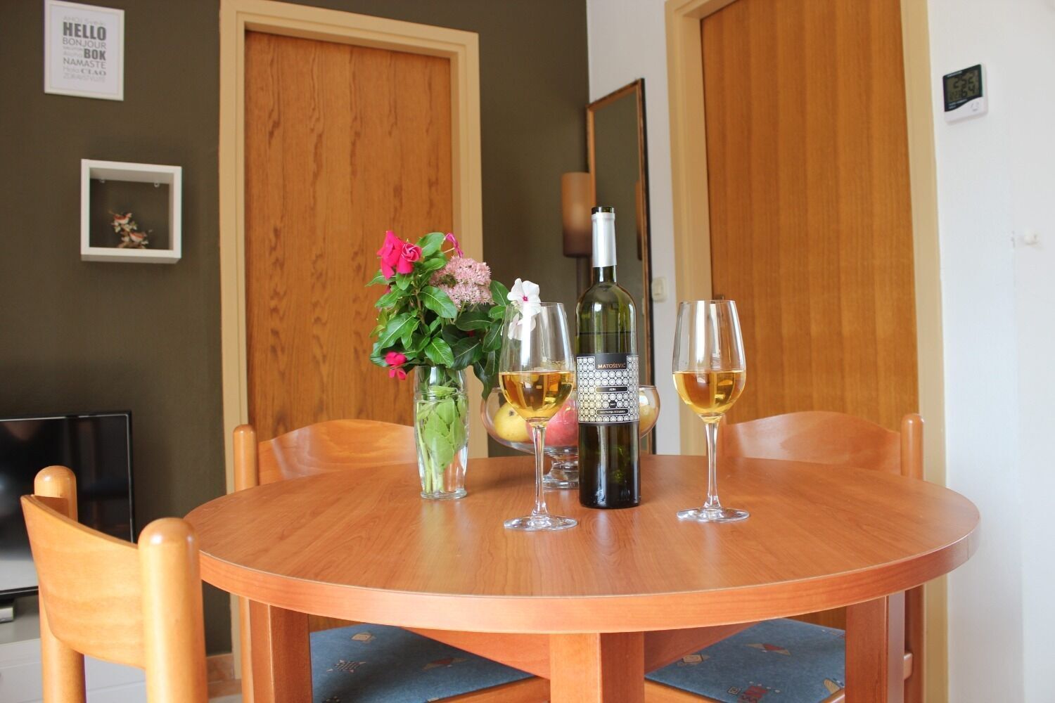 A round wooden table with a bottle of wine, two glasses of wine, and a vase with flowers.