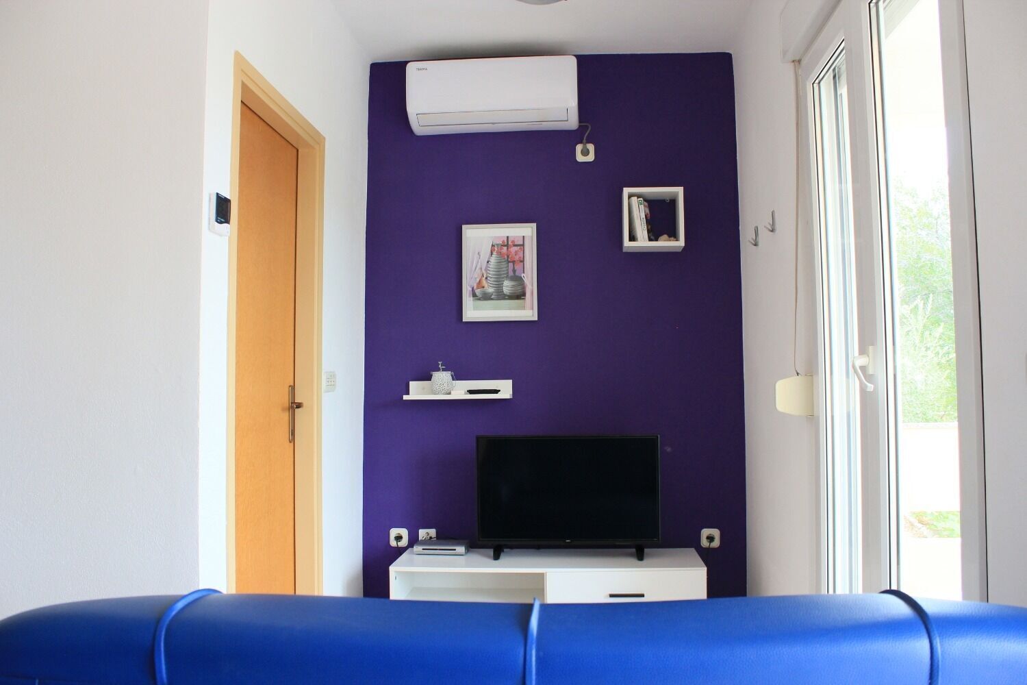 A room with a purple wall, a flat-screen TV on a white stand, a small shelf with decorative items, and a window with a view of greenery.