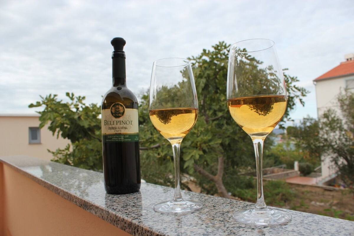 A bottle of Beli Pinot wine and two glasses on a balcony with a view of trees and a building.