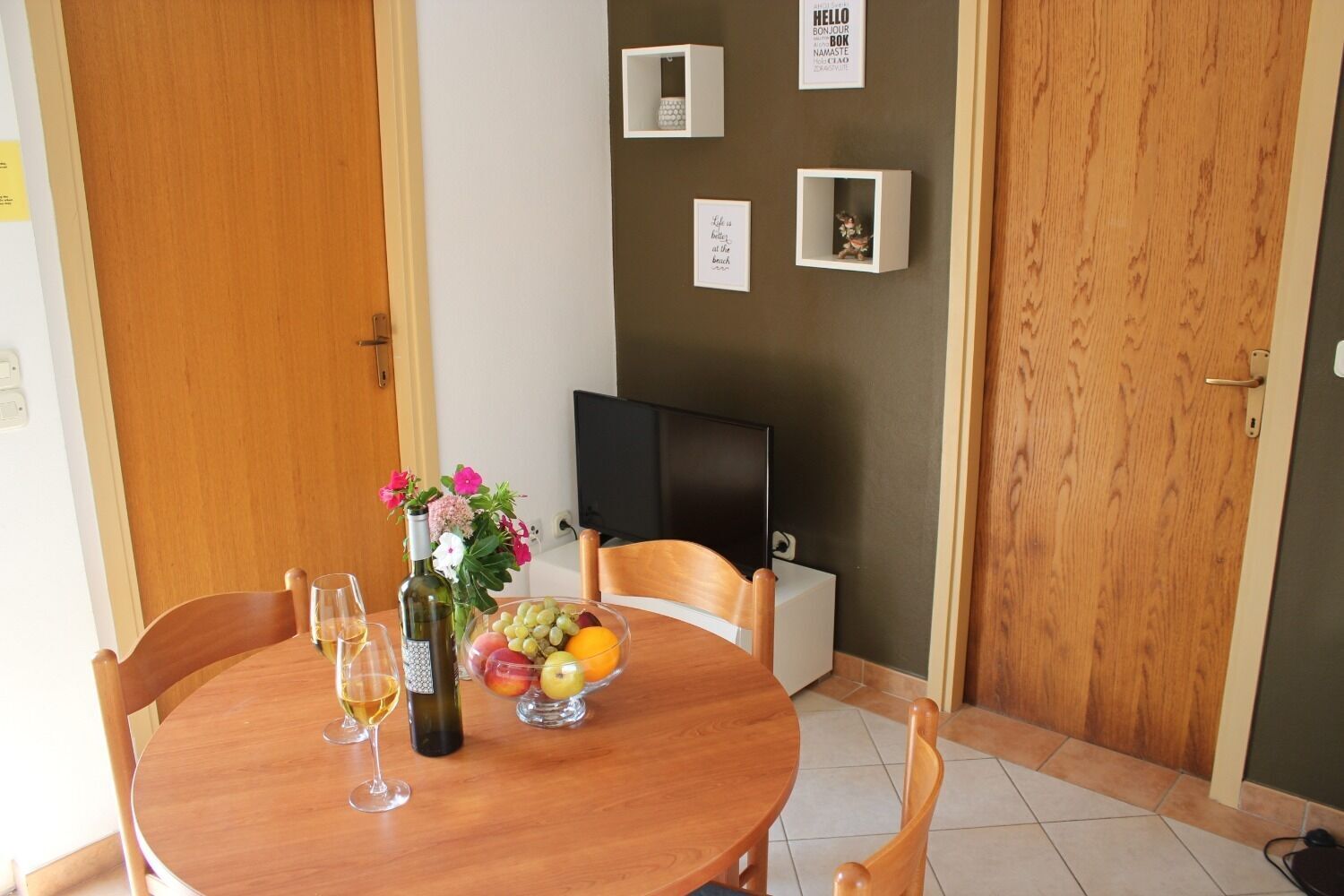 A dining area with a round wooden table, four chairs, a bottle of wine, a bowl of fruit, and a vase of flowers. There is a flat-screen TV mounted on the wall and a door with a wooden frame.
