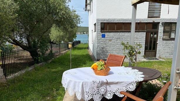 A table with a white tablecloth and a fruit bowl, a wooden chair, and a view of a body of water and a stone building. - Apartments Andy (Sukosan)