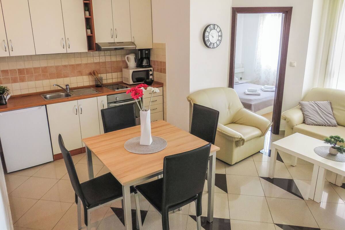 A modern kitchen with white cabinets, a coffee maker, and a microwave. There is a dining area with a table, chairs, and a vase with flowers. A sofa and a chair are in the living area.