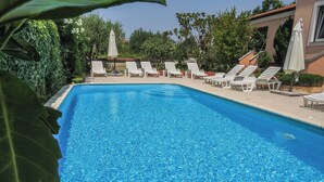 A clear swimming pool with white lounge chairs and umbrellas, surrounded by greenery and a building with a tiled roof. - Apartments Eddie (Zadar)