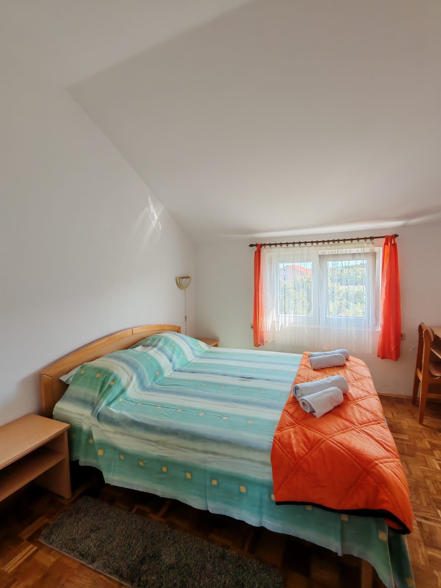 A bedroom with a bed, a nightstand, a chair, a window with curtains, and a view of the outside.