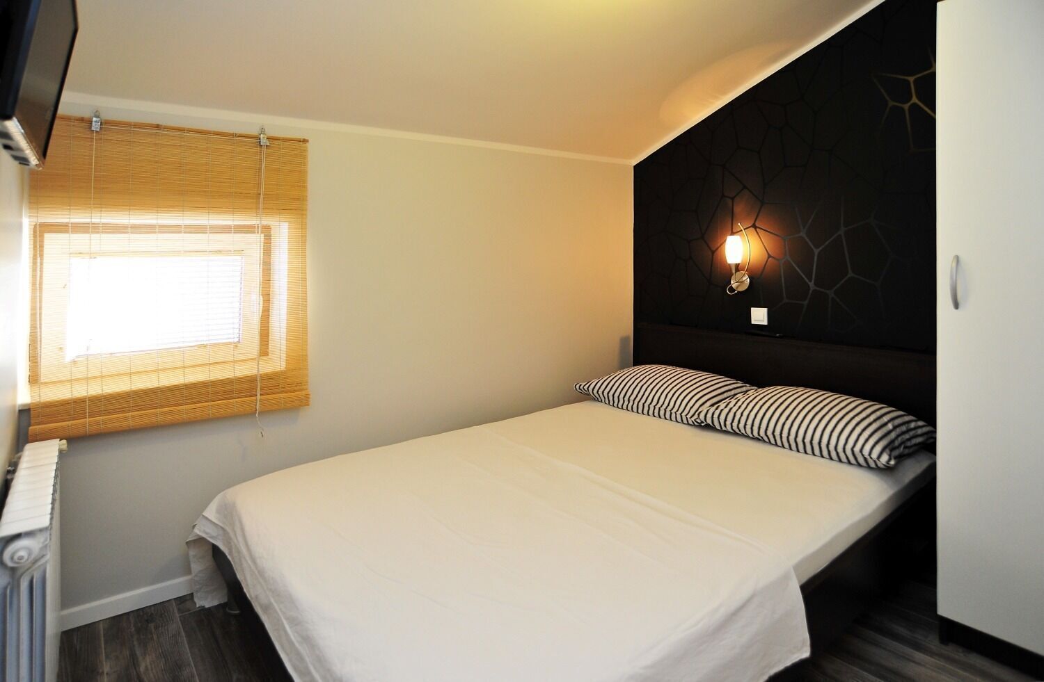 A single bed with a white mattress and striped pillows, a bedside table, a wall-mounted light, and a window with blinds.