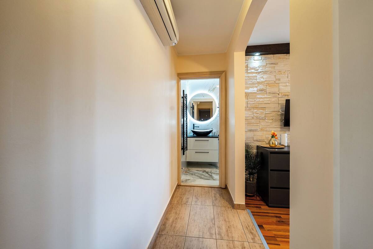 A hallway with a wooden floor, a kitchen area with a stone backsplash, and a modern light fixture.