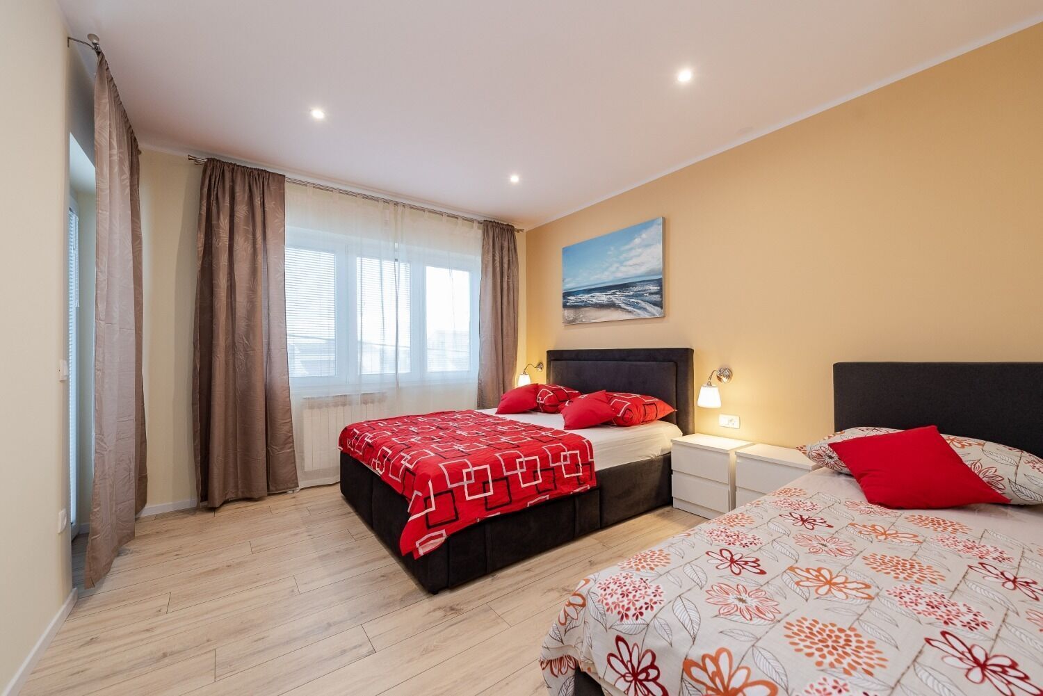 A bedroom with two beds, a large window with curtains, a painting on the wall, and a nightstand with a lamp.