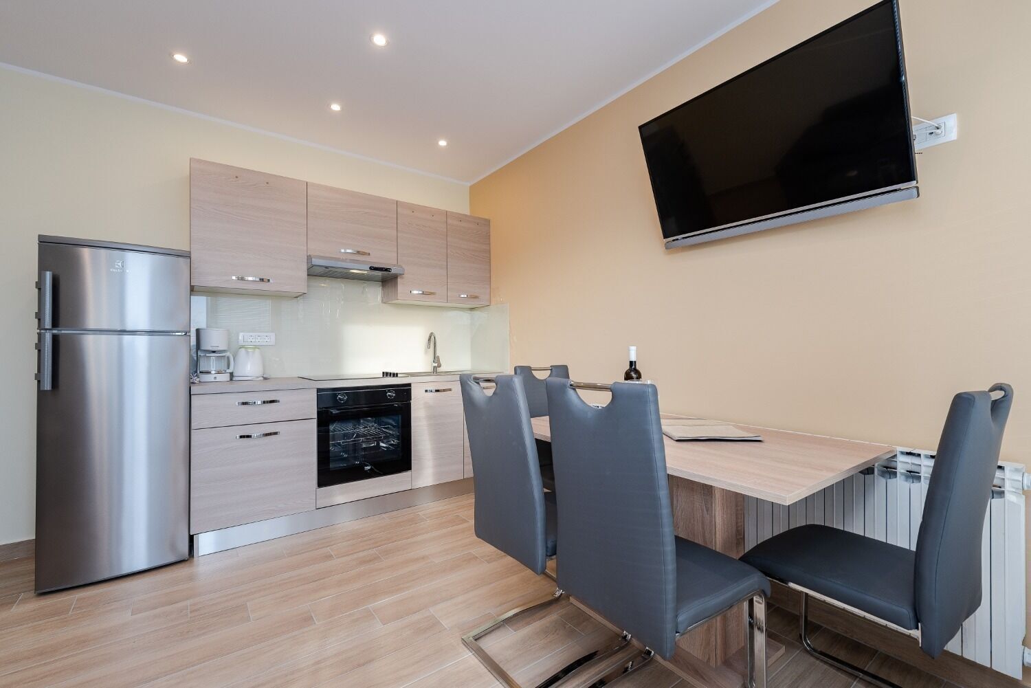 A modern kitchen with stainless steel appliances, a dining area with a table and chairs, and a mounted TV on the wall.