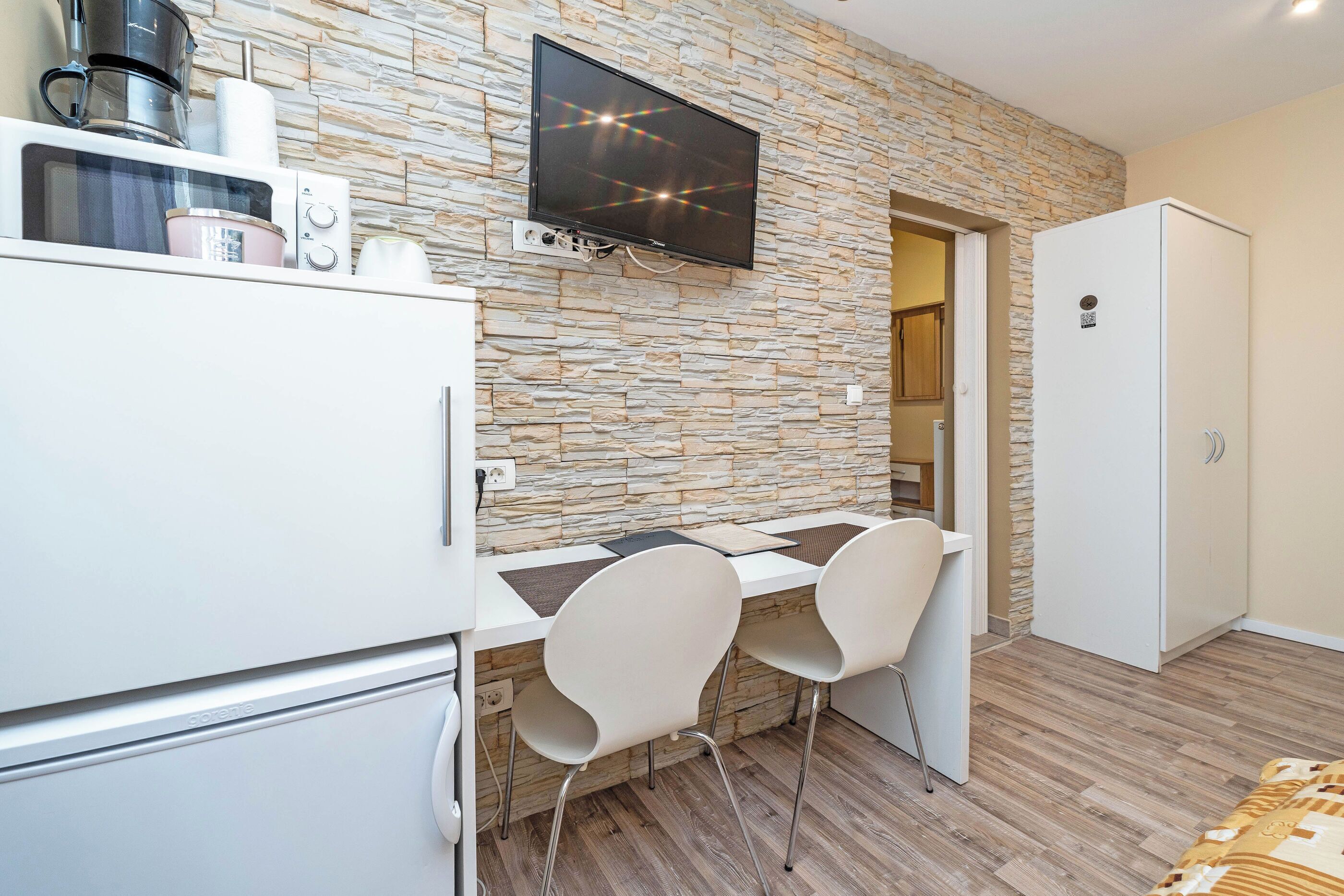 Una kitchenette equipada con microondas, cafetera y un televisor de pantalla plana montado en una pared de piedra. Hay dos sillas blancas y una mesa blanca frente a la kitchenette.