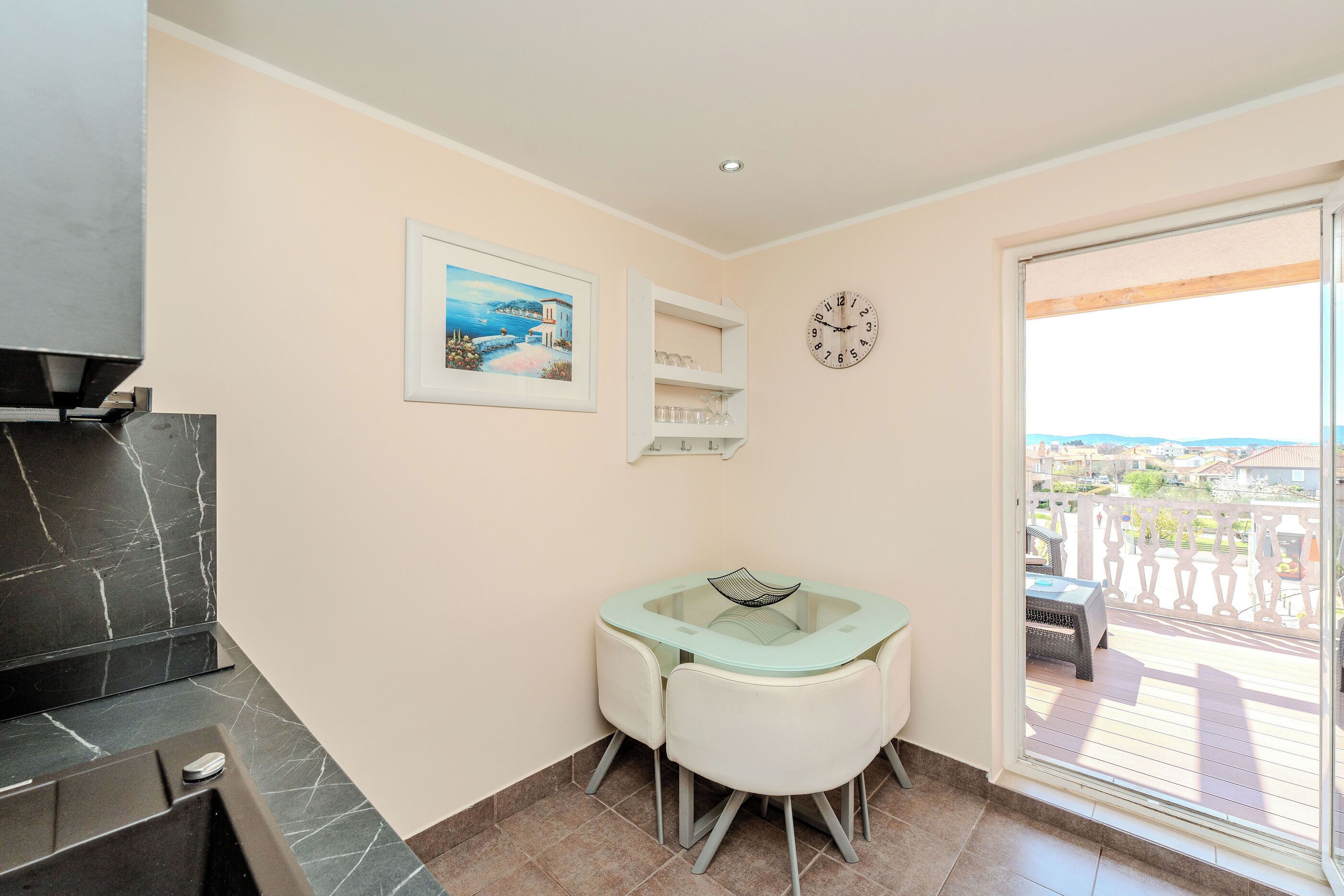A small room with a glass-top table, two chairs, a clock, a framed picture, and a view of the outside.