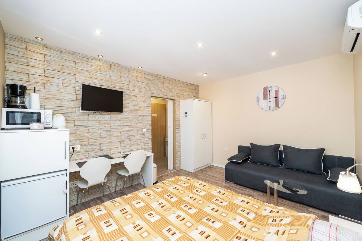 A modern room with a stone wall, a flat-screen TV, a microwave, a sofa, a coffee machine, and a bed with a patterned blanket.