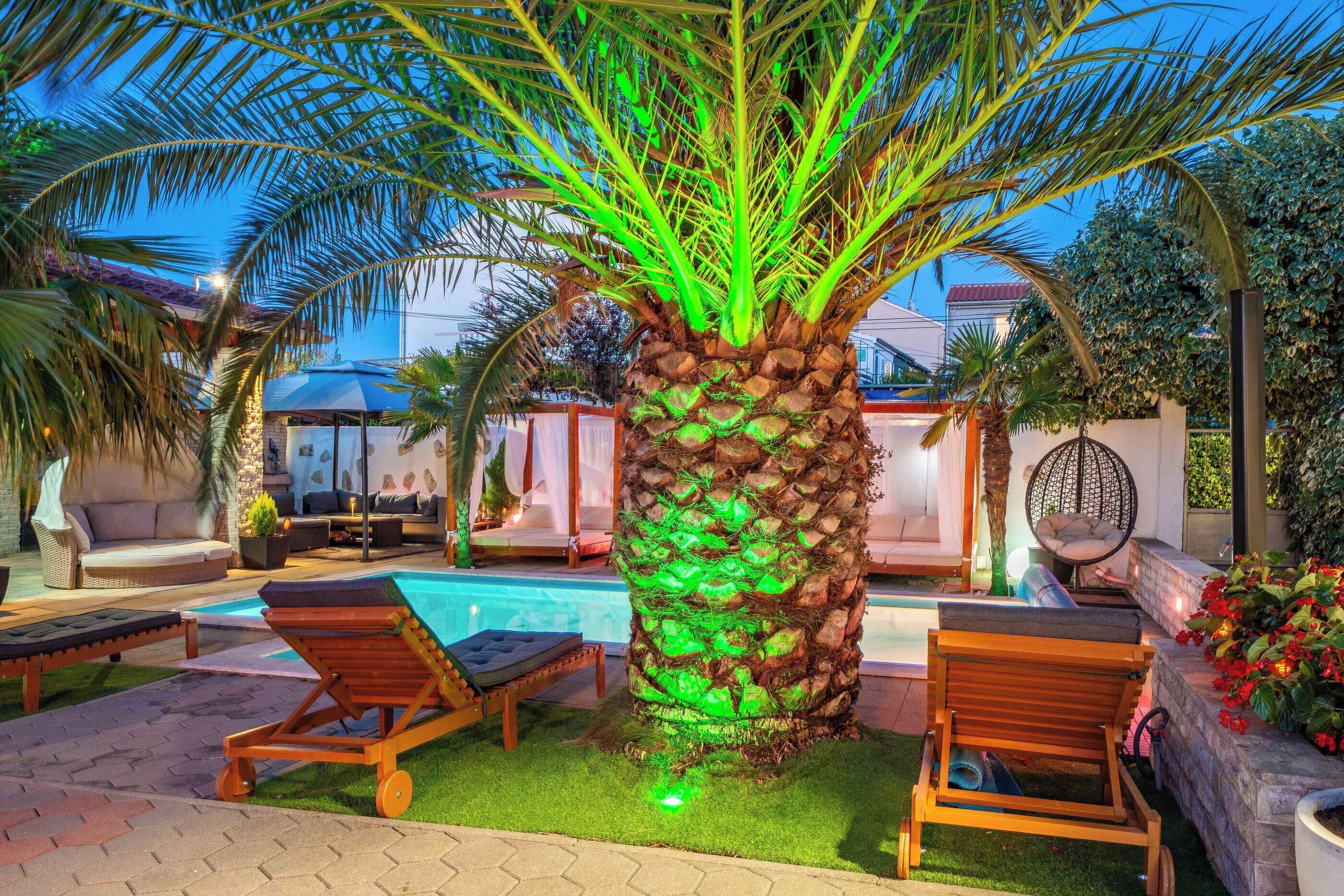 A poolside area with a palm tree illuminated by green lights, lounge chairs, and a small pool.