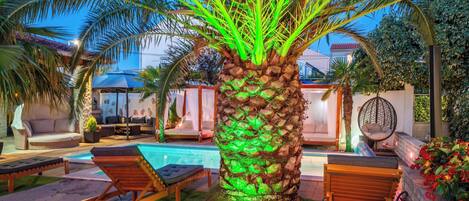 A poolside area with a palm tree illuminated by green lights, lounge chairs, and a small pool.