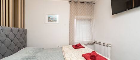 Et soveværelse med seng, radiator, et vindue med gardiner, et billede på væggen og et TV ophængt på væggen.