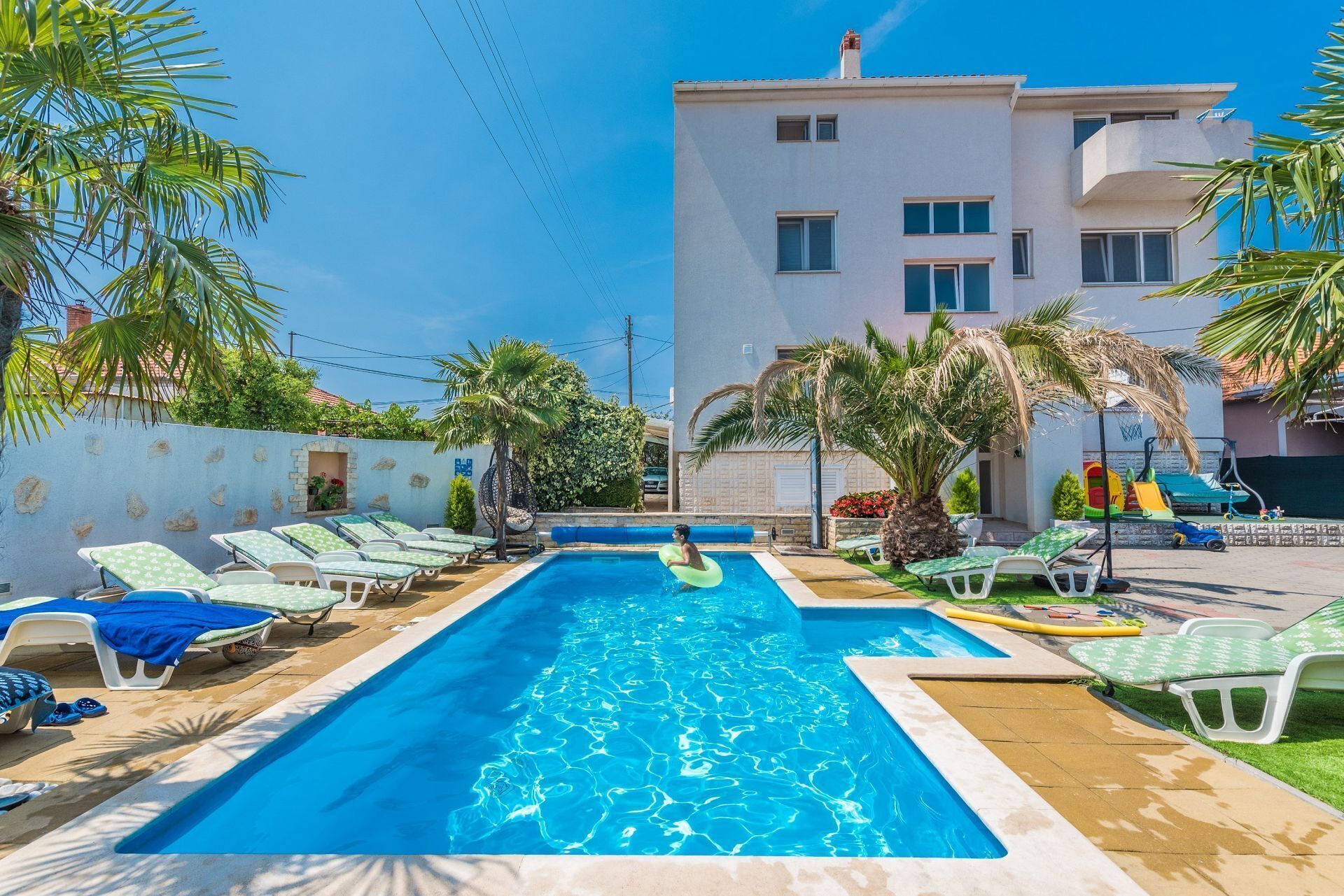 A swimming pool with lounge chairs and palm trees, surrounded by a building with multiple windows.