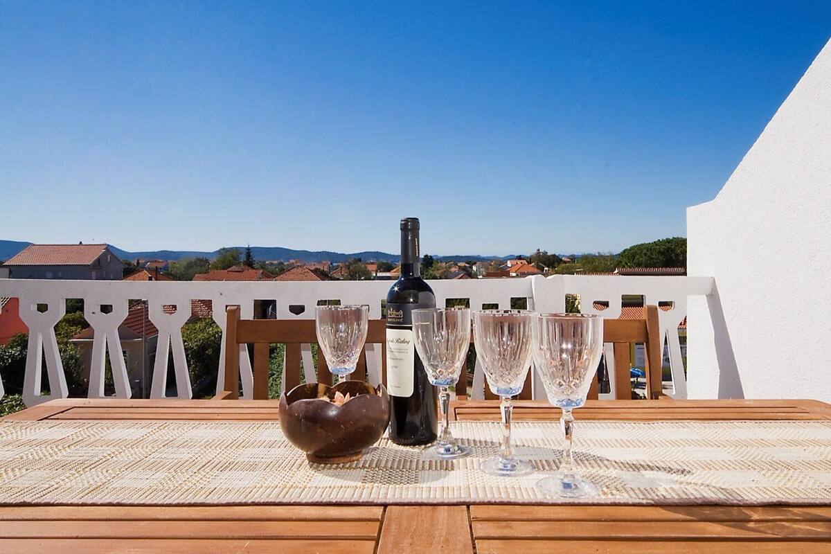 A wooden table with a bottle of wine and four wine glasses on a woven mat, set against a white fence with a scenic view of houses and mountains in the background.