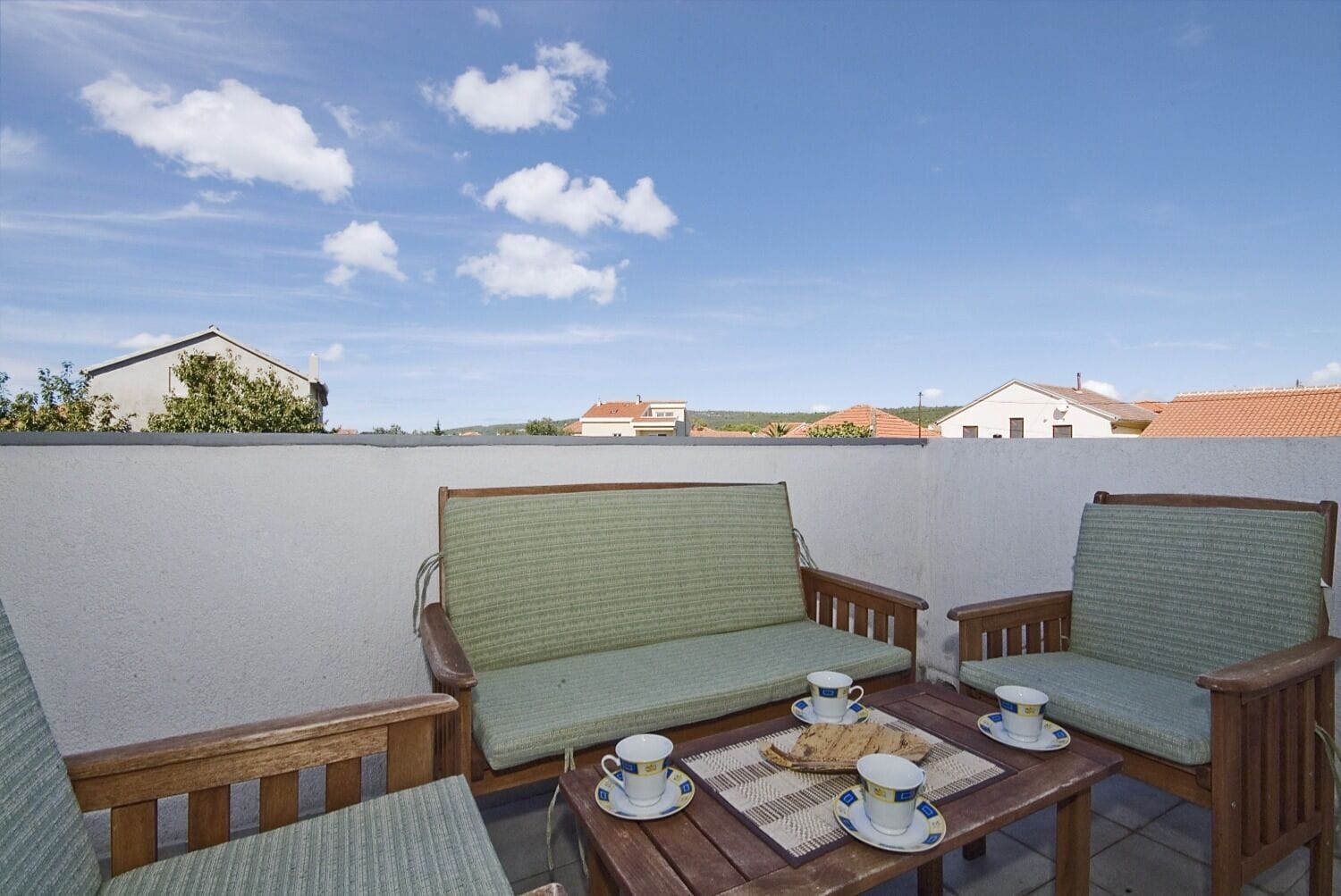 Pátio com dois sofás de madeira e uma mesa posta para chá, com vista para uma área residencial.