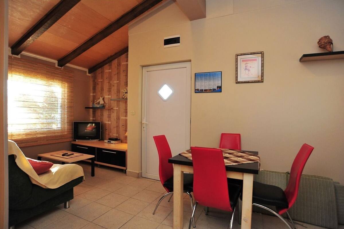 A living room with a TV, a sofa, a dining table with red chairs, and a window with blinds.