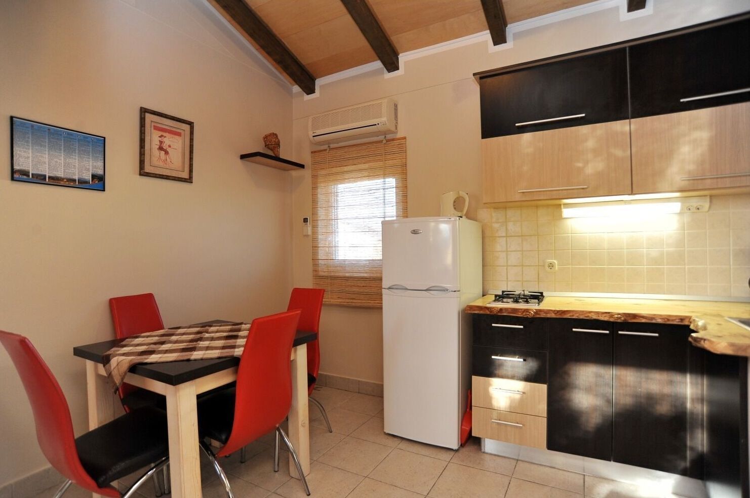 A compact kitchen with a refrigerator, a dining table with red chairs, and a window with blinds.