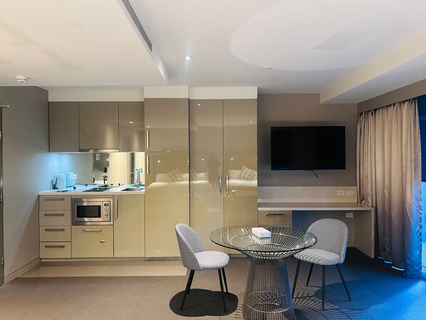 Een moderne hotelkamer met een kitchenette, een flatscreen televisie en twee geruite stoelen rond een glazen tafel.