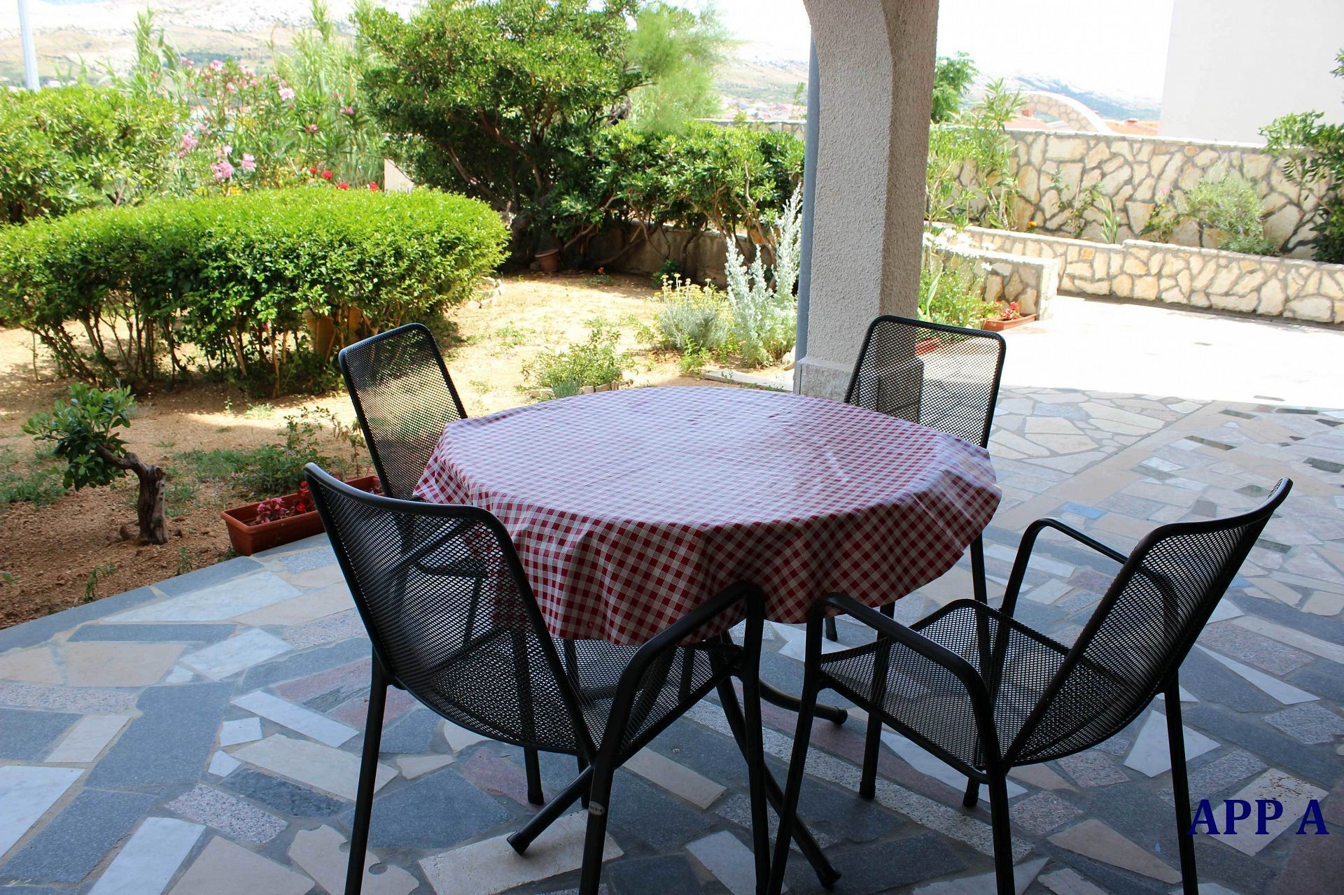 Mesa redonda con mantel a cuadros y cuatro sillas negras en un patio exterior con suelo de piedra y vegetación circundante.