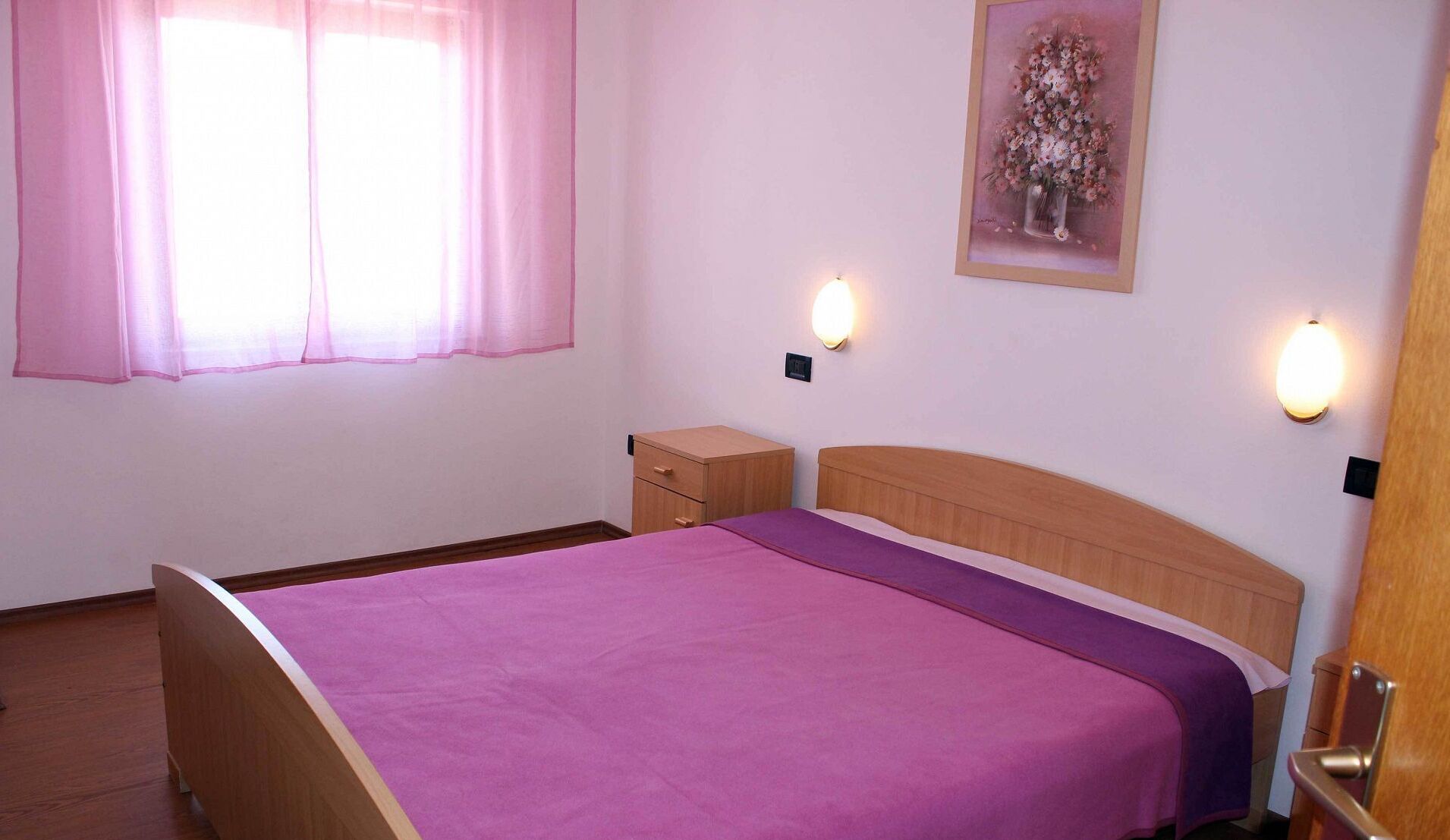 Une chambre à coucher comprenant un lit, une table de chevet, une tête de lit en bois, une fenêtre avec des rideaux roses et deux lampes fixées au mur.