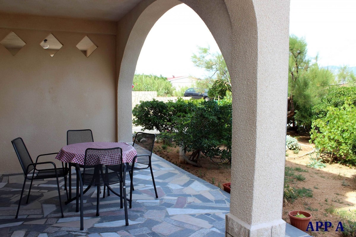 An outdoor patio with a table and chairs, a stone floor, and a view of a garden with trees and a car in the background.