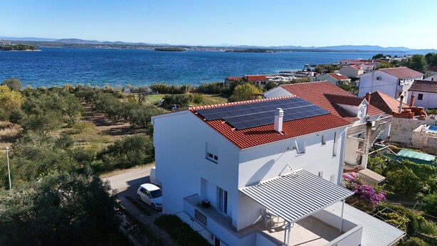 A modern white building with a red-tiled roof and solar panels, located near a body of water with a clear view of the sea and distant hills. - Apartments Mirko (Tkon)