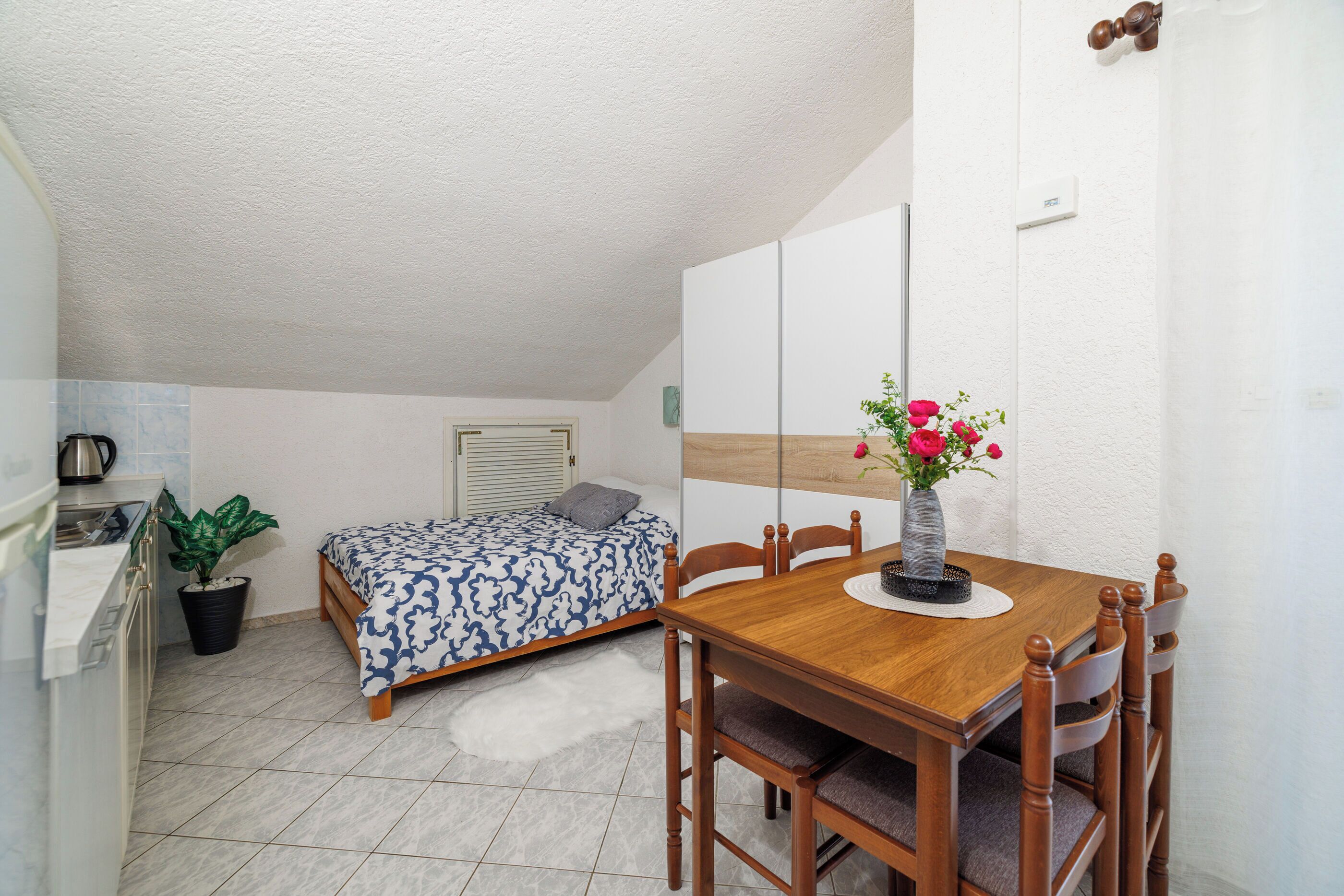 Una habitación pequeña con una cama, una mesa de comedor con sillas, una planta en maceta y una kitchenette.