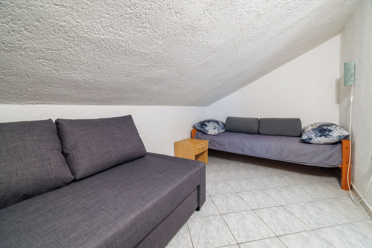 Una habitación pequeña y acogedora con un sofá gris y un banco a juego pegados a la pared, una mesita de noche de madera y techo blanco.