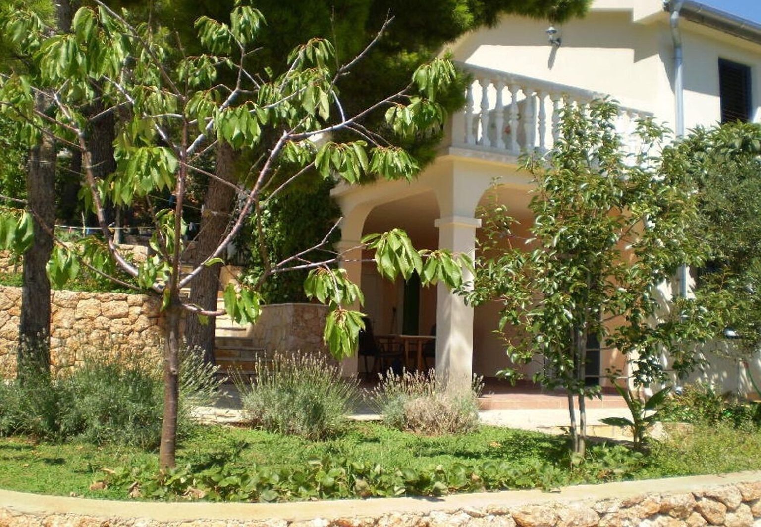 Uma casa com parede de pedra, varanda coberta e jardim com diversas plantas e árvores.