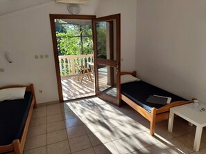 Ein kleiner, gut beleuchteter Raum mit zwei Holzbetten, einem weißen Tisch und einer Glastür, die auf einen Balkon mit einem Holzbett und Tisch führt.
