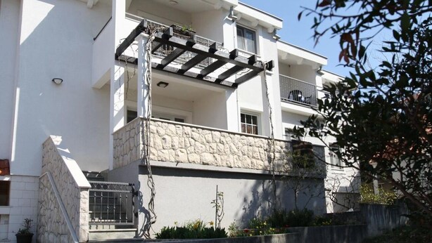 A two-story building with a stone facade, a balcony with a metal railing, and a small garden area with plants and flowers. - Apartments Mari (Zadar)
