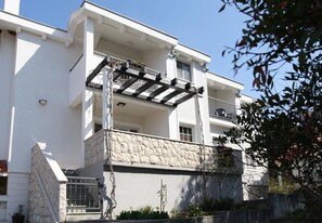 Ein zweistöckiges Gebäude mit einer Steinauffahrt, einem Balkon mit Metallgeländer und einem kleinen Gartenbereich mit Pflanzen und Blumen.