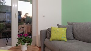 A grey sofa with a green pillow, a white pot with pink flowers, and a wooden table. - Apartments Mari (Zadar)