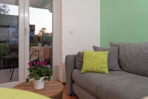 A grey sofa with a green pillow, a white pot with pink flowers, and a wooden table. - Apartments Mari (Zadar)