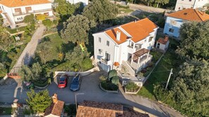 An aerial view of a residential area with a two-story house featuring a tiled roof, a driveway with parked cars, and surrounding greenery. - Apartments Mari (Zadar)