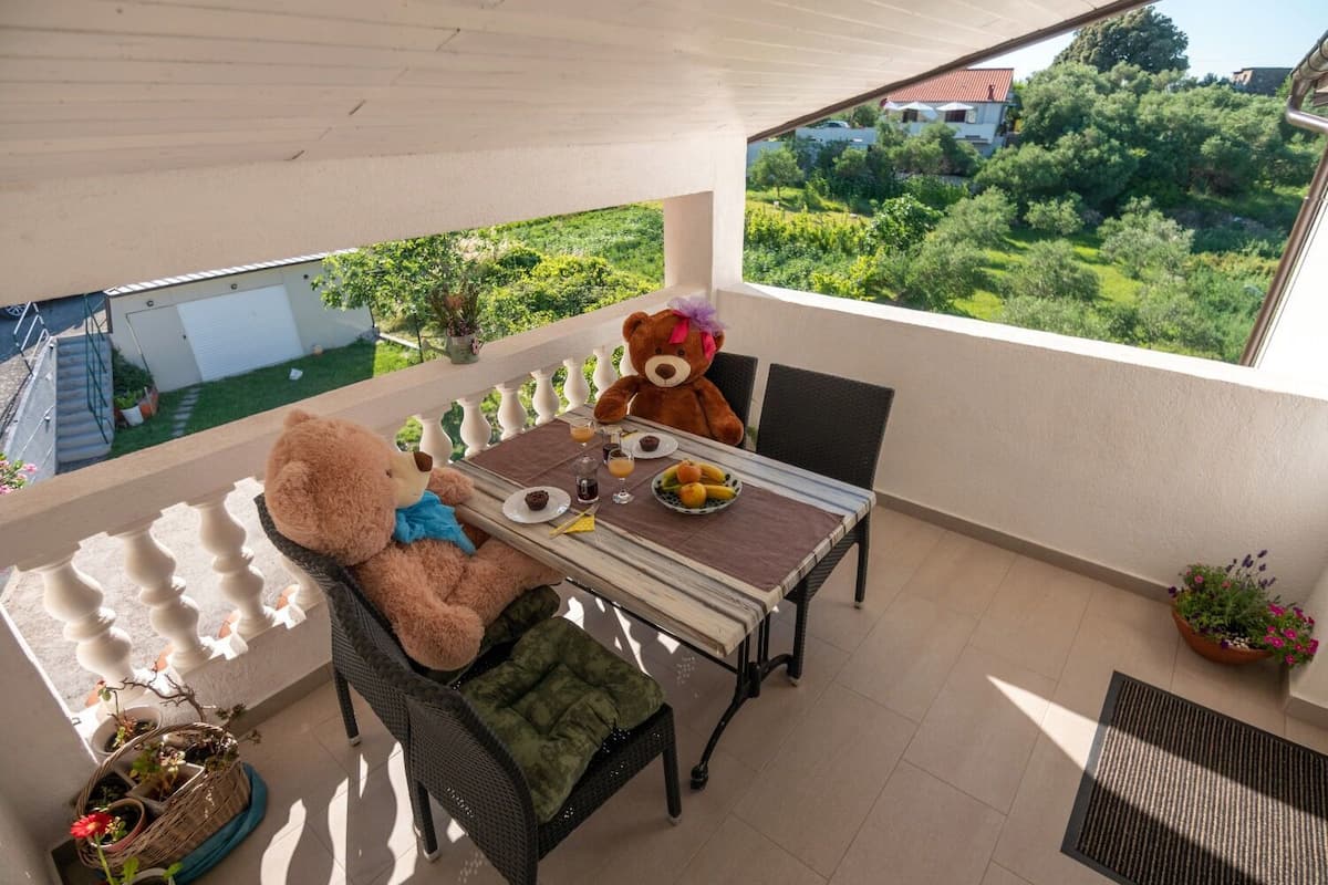 Un balcón con una mesa de comedor, dos sillas y dos osos de peluche. Hay plantas en macetas y se ve vegetación y edificios al fondo.