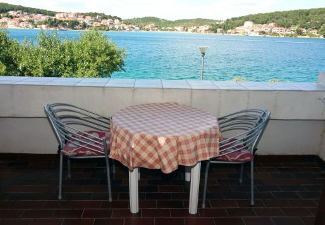 Une table recouverte d’une nappe à carreaux, avec deux chaises en métal dotées de coussins rouges, donnant sur une étendue d’eau et une ville lointaine.