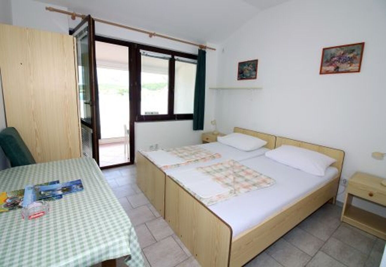 Ein Zimmer mit einem Bett, einem Tisch und einem Stuhl. Es gibt ein Fenster mit Vorhängen, ein an der Wand befestigtes Regal und einen kleinen Nachttisch.
