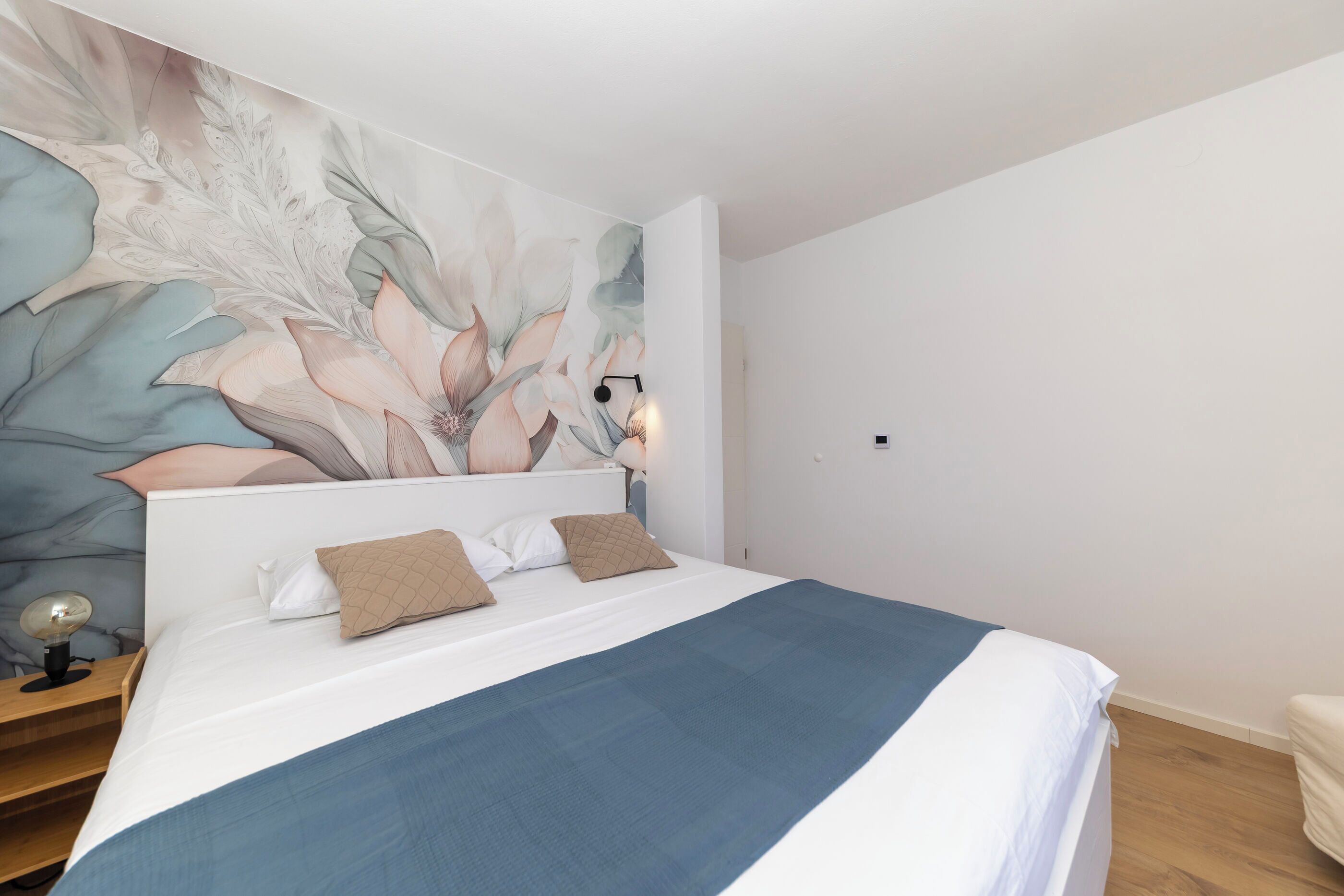 A bedroom with a bed, a nightstand, a wall-mounted lamp, and a large floral mural.