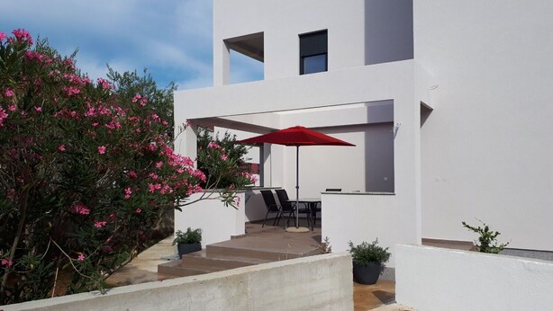 A modern white building with a patio area featuring a red umbrella, outdoor seating, and potted plants. - Apartments Mari (Nin)