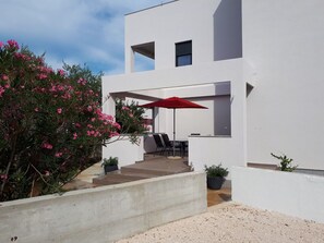 Ein modernes, weißes Gebäude mit einer Terrassenfläche, die einen roten Sonnenschirm, Sitzgelegenheiten im Freien und Topfpflanzen zeigt.