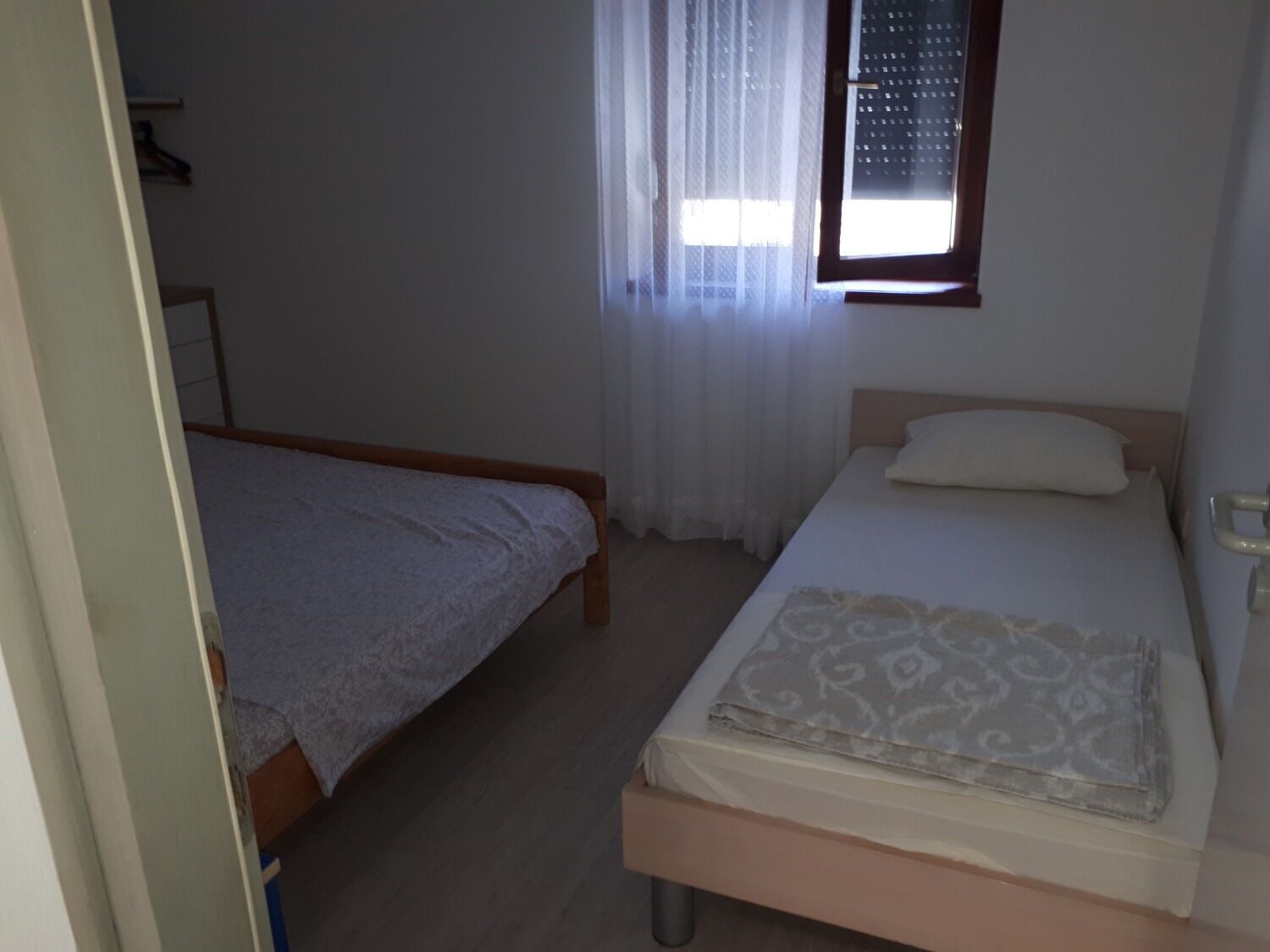 A small, simple bedroom with two single beds, a window with curtains, and a plain wall.