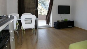 Een compacte kamer met een kitchenette, een eethoek met een witte stoel, een televisie aan de muur en een raam met gordijnen.